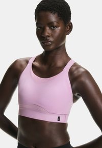 Brassière de sport rose clair à la texture lisse, avec des bretelles larges, un col arrondi et un logo noir en accent sur la bande inférieure.