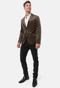 Mørk oliven fløyelsblazer med spiss krage, enkel knappelukking, to frontlommer, båret over en hvit skjorte og svarte bukser. Svarte sko.