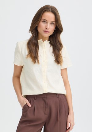Femme aux longs cheveux bruns portant un chemisier crème à manches courtes boutonné et un pantalon marron taille haute, une main dans la poche.