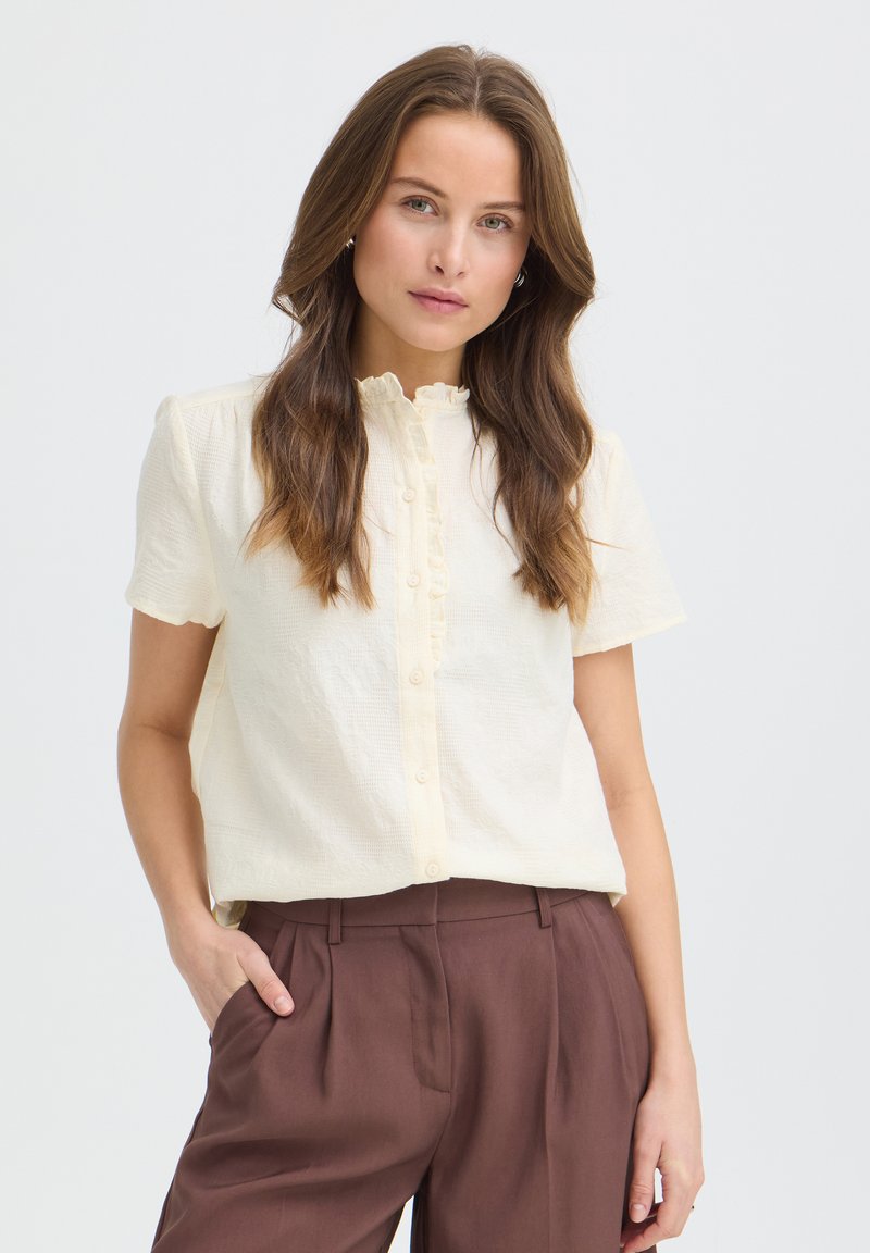Femme aux longs cheveux bruns portant un chemisier crème à manches courtes boutonné et un pantalon marron taille haute, une main dans la poche.