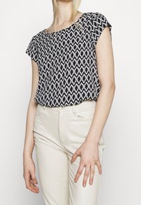Top a maniche corte con pattern bianco e nero, dal taglio morbido, abbinato a pantaloni beige attillati. Tessuto testurizzato con design geometrico.