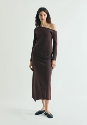 Kvinde med langt mørkt hår iført en mørkebrun off-shoulder langærmet top og matchende asymmetrisk midi-nederdel med sorte slip-on sko.