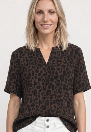 Femme souriante aux cheveux blonds mi-longs, portant un haut marron à manches courtes avec des taches léopard noires et un pantalon blanc.