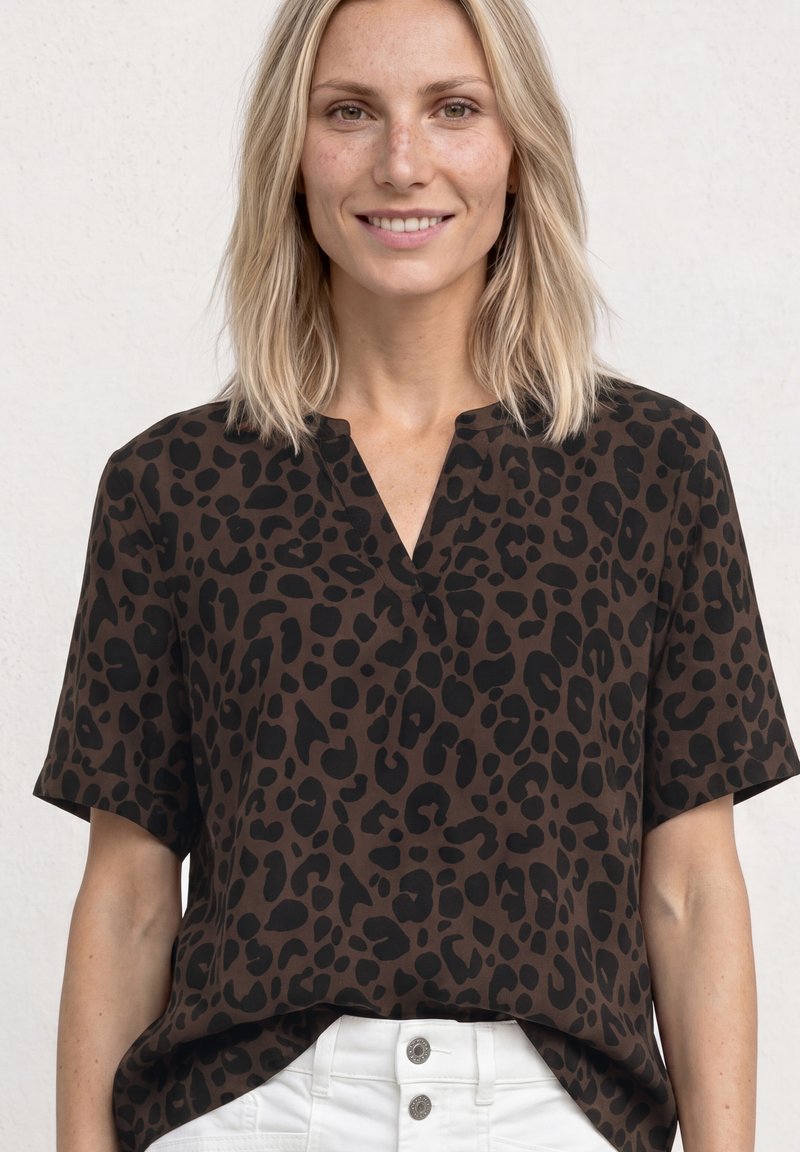 Femme souriante aux cheveux blonds mi-longs, portant un haut marron à manches courtes avec des taches léopard noires et un pantalon blanc.