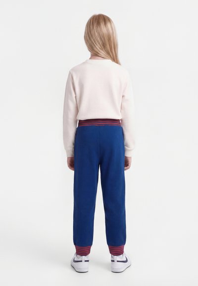 Dieťa s dlhými blond vlasmi stojí chrbtom, oblečené v svetloružovom svetri, modrých teplákoch s červenými pruhmi a bielych teniskách.