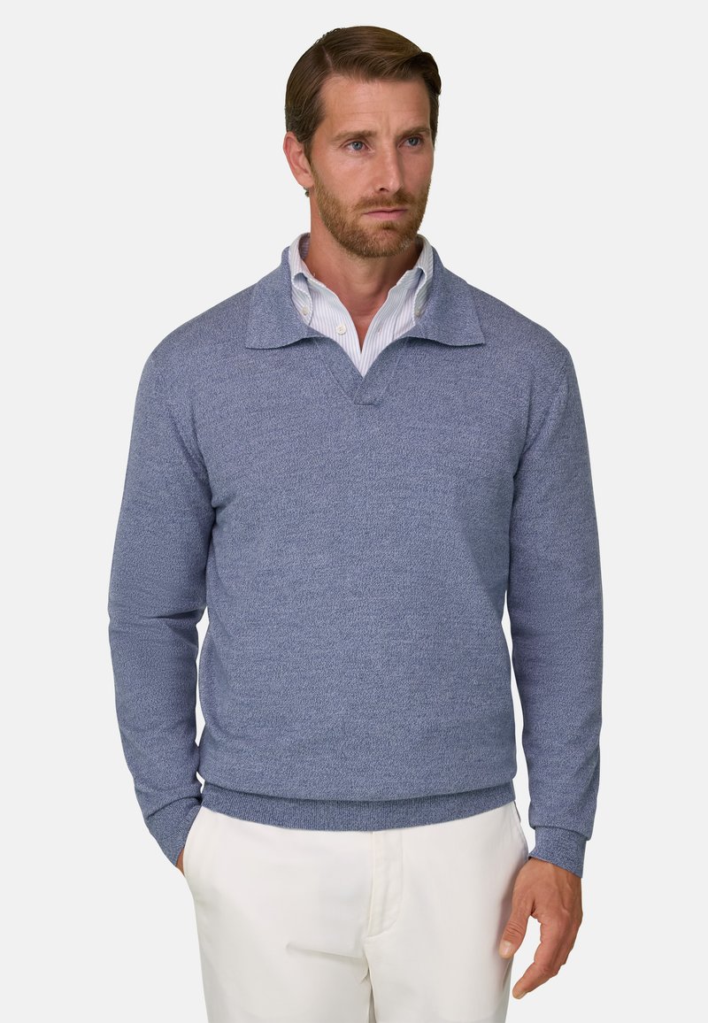 Homme portant un pull bleu à manches longues avec un col sur une chemise blanche et un pantalon blanc, debout devant un fond uni.