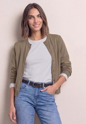 Femme aux cheveux bruns mi-longs, portant une chemise blanche, un cardigan vert olive, un jean bleu et une ceinture noire, souriante avec une main dans la poche.
