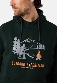 Felpa verde con un grafiche di una persona che pesca, circondata da alberi, montagne e un falò. "ESPEDIZIONE ALL'APERTO" stampato in arancione.