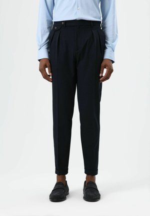 Pantalon plissé bleu marine à coupe fuselée, confectionné dans un tissu léger. Il comporte des poches avant, un bouton latéral et des ourlets retroussés.