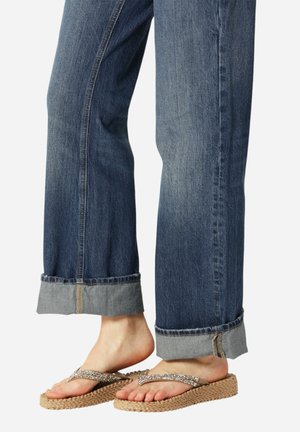 Bare feet wearing sparkly strap woven sole slippers paired with blue cuffed denim jeans.