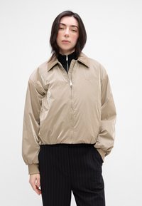 Beige nylon bomber jacket with a front zipper, oversized fit, and large collar. Paired with black pinstriped pants and a black top.