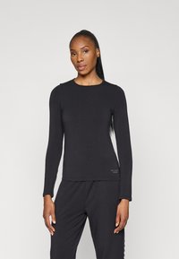 Garšām piedurknēm melna blūze gludā audumā, ar apaļu kaklu un smalku zīmolu uz apakšmalas. Kombinēta ar saskaņojošām melnām biksēm.