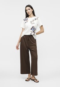 Kvinde iført hvid T-shirt med blå blomstermønster, løse brune bukser og sorte sandaler, stående med den ene hånd på hoften.