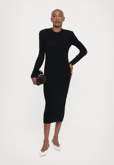 Rochie neagră cu dungi, cu mânecă lungă, siluetă ajustată, decolteu rotund și decupaje laterale. Modelul ține o geantă mică, rotundă, de culoare neagră.