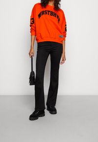 Sudadera naranja con letras y acentos en negro, combinada con jeans negros de bota acampanada y zapatillas negras gruesas, sostenida con un pequeño bolso negro.