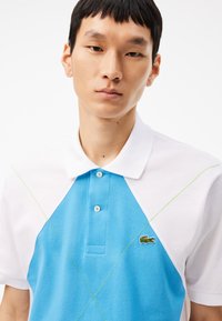 Polo con un frente azul y mangas blancas, de algodón texturizado, con una tapeta de tres botones, costuras de acento verdes y un pequeño logo.