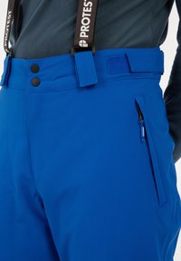 Modré snehové nohavice s hladkým povrchom, nastaviteľným pásom, dvoma čiernymi gombíkmi a bočnou zipsovou kapsou. Majú značkové traky.