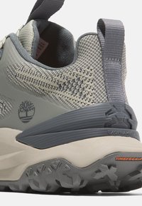 Zapatilla gris y beige con una parte superior de punto texturizado, talón reforzado y suela de goma. Las características incluyen el logo de Timberland y una pestaña de agarre.