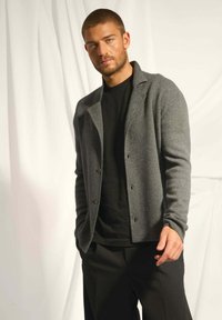 Grå ullkofta med slagkrage, två knappar och långa ärmar, lagd över en svart t-shirt, matchad med svarta byxor.