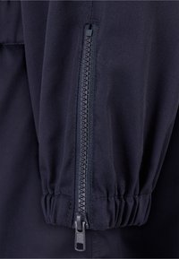 Gros plan sur une manche de veste bleu marine foncé avec un poignet élastique et une fermeture éclair noire verticale.