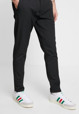 Person iført sorte slim-fit bukser med hænderne i lommerne og hvide sneakers med grønne og røde striber på en ensfarvet baggrund.