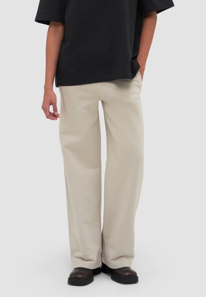 Osoba ubrana w beżowe spodnie o szerokich nogawkach, czarną oversize koszulę i czarne buty, stojąca na jednolitym jasnoszarym tle.