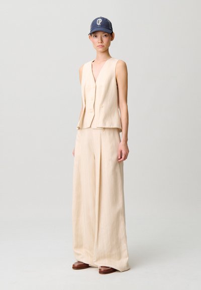 Gilet beige a righe con scollo a V e bottoni, abbinato a pantaloni a gamba larga coordinati. Completato da un berretto blu con logo bianco.