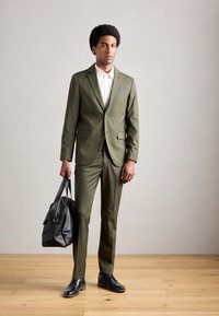 Hombre con traje verde olivo y camisa blanca sosteniendo un bolso de cuero negro, de pie sobre un suelo de madera frente a una pared gris claro lisa.