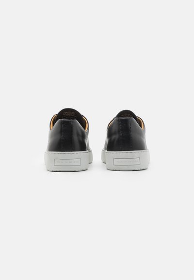 Sapatilhas slip-on de couro preto com uma sola de borracha branca, exibindo uma textura suave e uma marcação discreta no calcanhar.