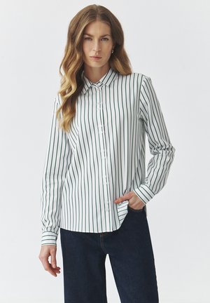 Camicetta bianca a maniche lunghe con righe verticali verdi, dotata di colletto a punta e maniche con polsini. Indossata con jeans scuri in denim.