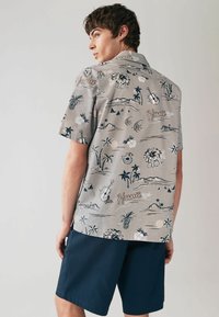 Rövid ujjú ing világosszürke alapszínen, amely hawaii témájú grafikákat tartalmaz, beleértve pálmafákat, hegyeket és különböző sötétebb színű motívumokat.