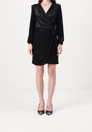 Kobieta ubrana w czarną, kopertową sukienkę z długim rękawem z subtelnym wzorem oraz czarne szpilki z ostrym noskiem, stojąca na białym tle.