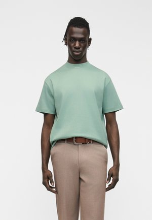 Άνδρας με σκουρόχρωμο δέρμα και πλεξούδες, ντυμένος με πουκάμισο κοντομάνικο σε μέντα πράσινο και ανοιχτό καφέ παντελόνι, στέκεται μπροστά σε απλό λευκό φόντο.