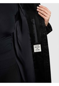 Melnā žakete no iekšpuses ar gludu materiālu, rāvējslēdzēja aizdari un etiķeti ar uzrakstu "Siltas apskāviens no auduma."