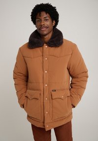 Casaco acolchoado castanho com gola de pelo escuro, botões de pressão e dois bolsos frontais. Tecido texturado com elementos de design acolchoados.