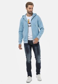 Un sweat à capuche zippé bleu clair avec un cordon de serrage, associé à un t-shirt graphique blanc et un jean foncé. Des baskets blanches complètent l'ensemble.