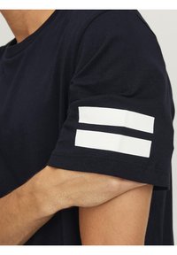 Marinblå t-shirt med korta ärmar som har två horisontella vita ränder på vänstra ärmen. Tillverkad av ett mjukt, texturerat tyg.