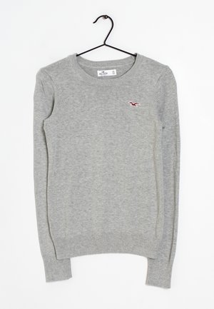 Pull en maille gris clair à manches longues avec col rond et petit logo d'oiseau rouge sur la poitrine gauche, suspendu à un cintre noir.