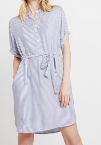 Sukienka w paski w kolorze białym i granatowym, z krótkimi rękawami, zapięciem na guziki z przodu, paskiem w talii oraz kieszeniami bocznymi. Wykonana z miękkiego materiału, luźny krój.