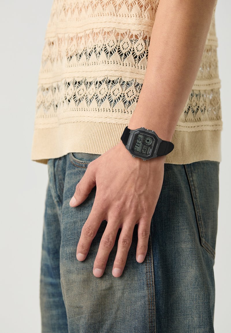 Montre numérique avec un bracelet en plastique noir, cadran rectangulaire affichant l'heure et les fonctions, portée sur une main contre un tricot texturé de couleur claire et un jean.