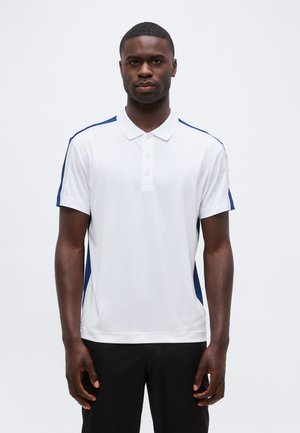 Homme portant un polo blanc à manches courtes avec des bandes bleues sur les côtés et un pantalon noir, debout face à l'avant sur un fond uni.