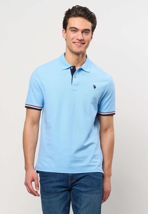 Fiatal férfi mosolyog, világoskék rövidujjú galléros pólót visel csíkos ujj részletekkel és sötétkék farmert, egyszínű háttér előtt áll.