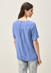 Blusa a maniche corte di colore azzurro chiaro con scollo rotondo, dettagli a pieghe nella parte posteriore e orlo curvo. Tessuto dalla superficie liscia, vestibilità rilassata.