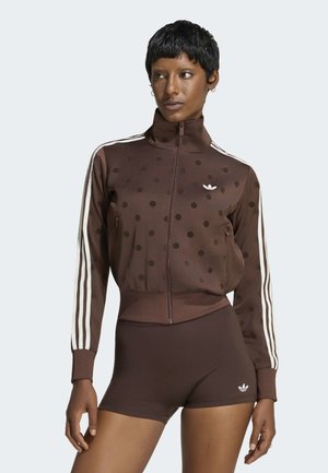 Kvinde iført brun polkaprikket jakke med lynlås og hvide striber på ærmerne samt matchende brune shorts med logo på venstre ben.
