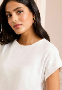 Haut blanc à manches courtes avec un col rond. Le tissu semble lisse et léger. Des boucles d'oreilles en forme de croissant doré sont portées.
