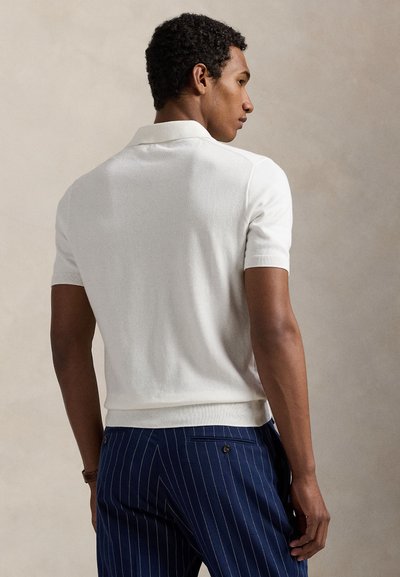 Háttal álló férfi, fehér rövid ujjú galléros pólót és tengerészkék sávos nadrágot visel, semleges háttér előtt.
