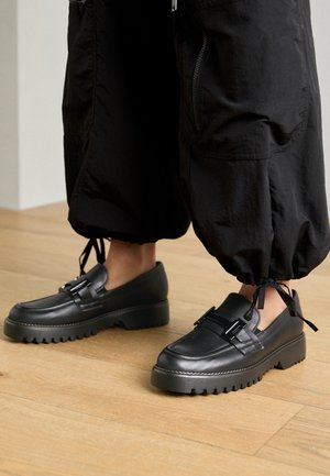 Chaussures noires en cuir avec une semelle épaisse et rainurée ainsi qu'une tige lisse, dotées de deux boucles métalliques et d'une forme arrondie à l'avant.