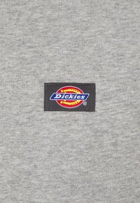 Tessuto grigio con finitura testurizzata che presenta una toppa rettangolare nera. La toppa mostra il logo "Dickies" nei colori blu, rosso e giallo.