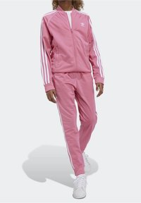 Różowy dres wykonany z miękkiego materiału, składający się z kurtki na zamek z białymi paskami i logo, w zestawie z dopasowanymi spodniami i białymi butami.
