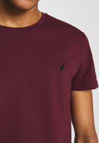 Bordo pamučna majica s okruglim izrezom, kratkim rukavima i malim crnim izvezenim logom ptice na lijevoj strani prsiju.